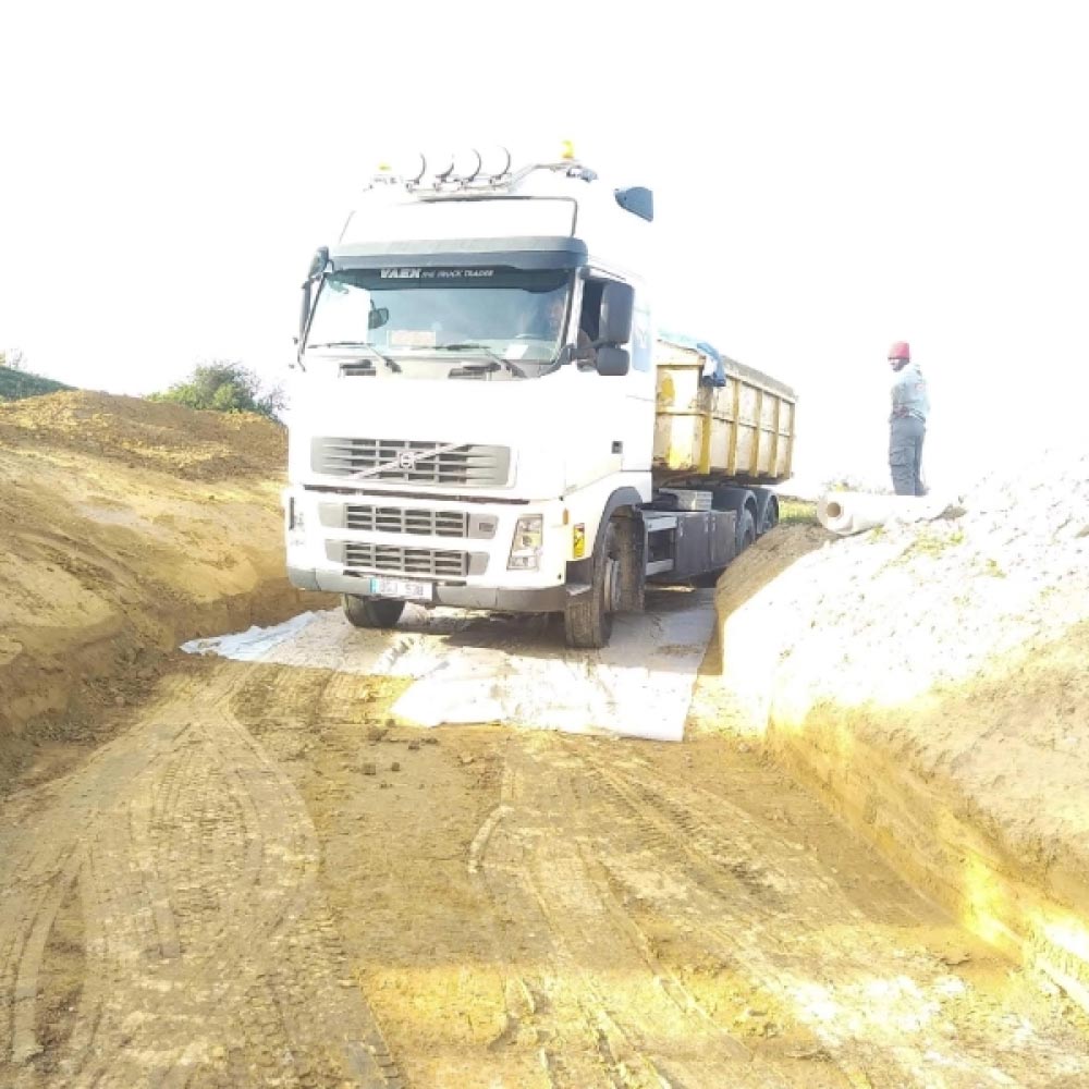 Camion Volvo blanc avec benne jaune traversant une tranchée boueuse sur des nattes de stabilisation, un ouvrier observe.