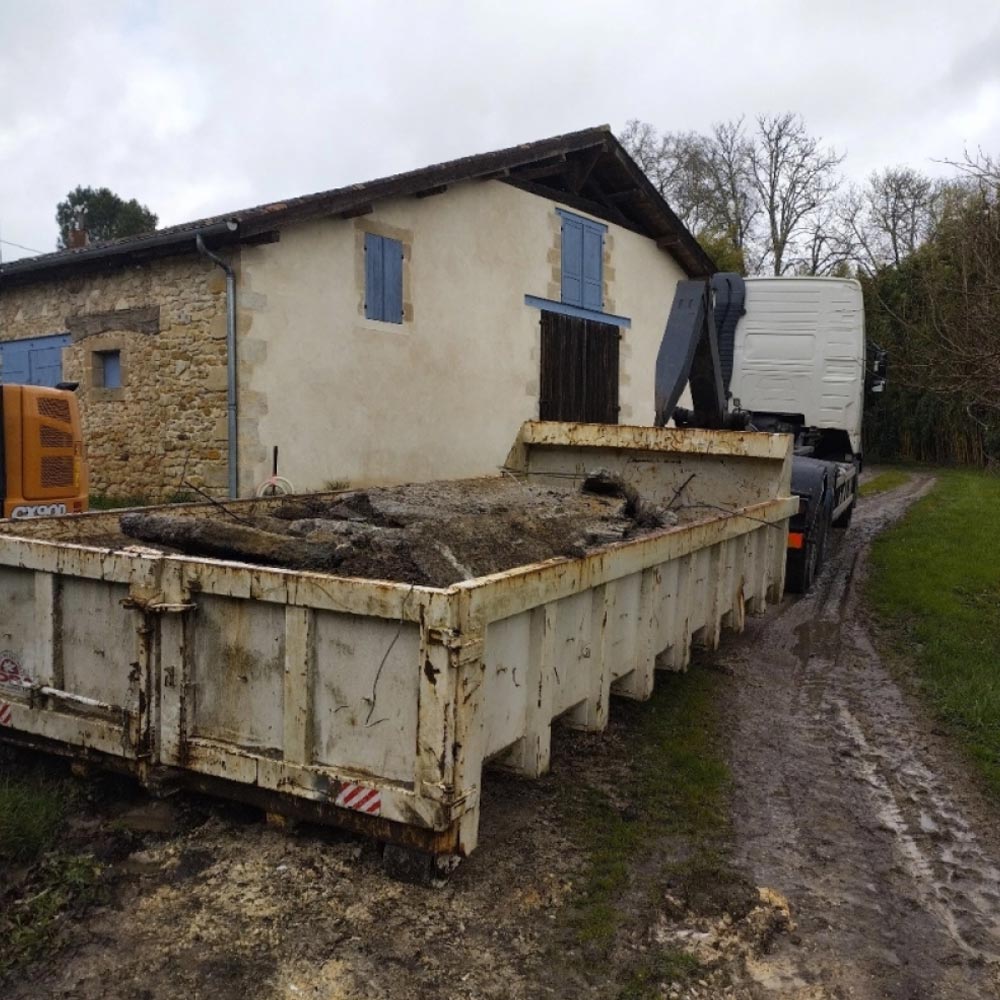 Camion avec benne remplie de débris et terre devant une vieille ferme en pierre et crépi, volets bleus, chemin boueux.